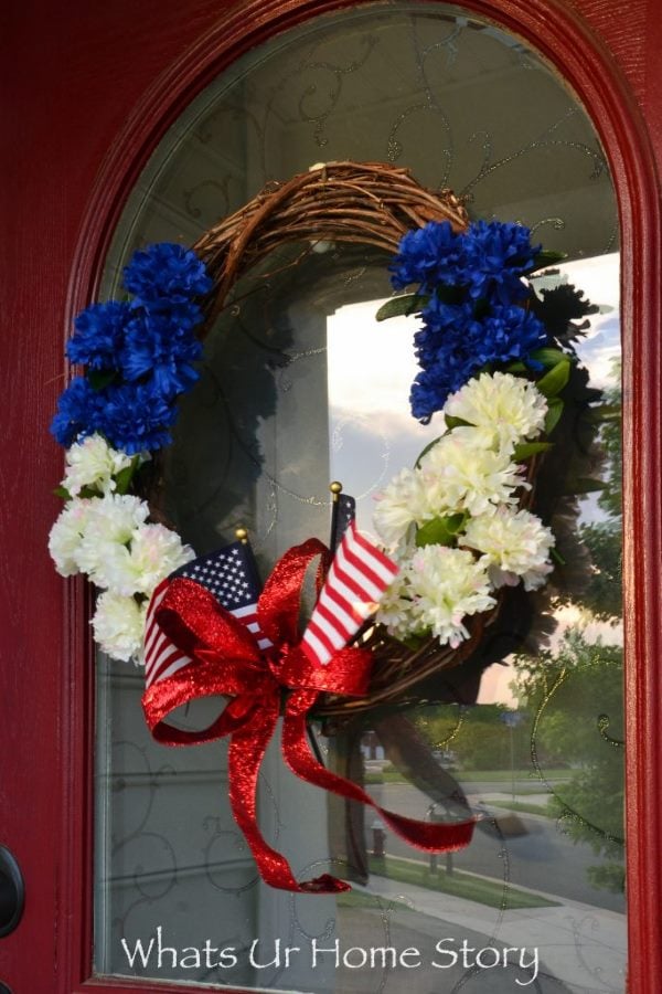 Patriotic grapevine wreath with blue and white flowers, American flags, and red glitter ribbon bow