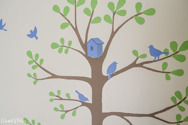 Bright children's bedroom with a hand-painted tree mural featuring birds, leaves, and colorful nature-inspired details.