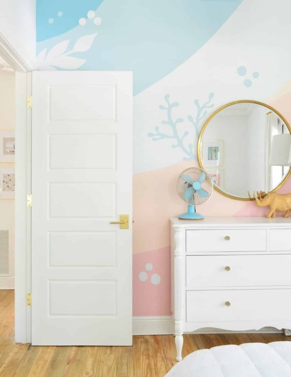 Kids' bedroom with a playful geometric wall mural painted in soft blue, peach, pink, and cream color blocks.