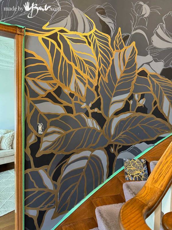 Dark accent wall painted with oversized botanical leaves outlined in metallic gold, creating a dramatic floral wall mural.