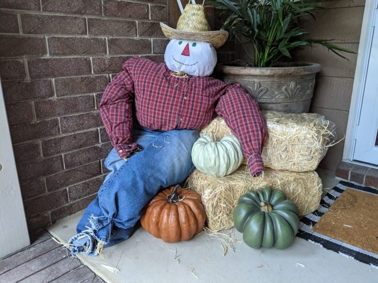 Simple homemade scarecrow with a smiling fabric face, plaid shirt, jeans, straw hat, and rubber boots.