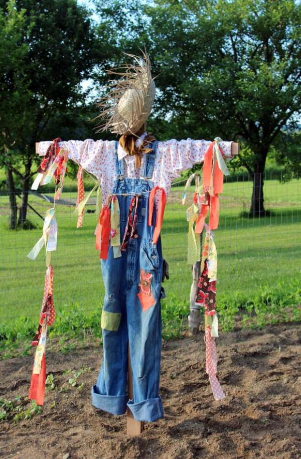 Classic DIY scarecrow in a plaid shirt and jeans standing beside raised garden beds.